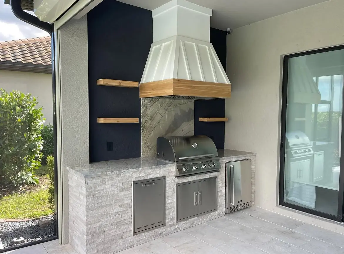 Custom site-built outdoor kitchen with stone finish, wood-trimmed hood and floating shelves